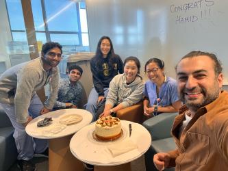 The Doris Wang lab sits around two small coffee tables getting ready to cut into a cake while Hamid takes a selfie. The white board behind them reads, Congrats Hamid!!!